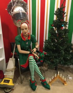 A bald mannequin dressed in christmas attire seated next to a vaccum